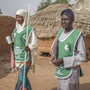 Photo of a woman and man walking through a rural Nigerian village. Text reads I wantto break the transmission explains Maryam a community drug distributor in Nigeria