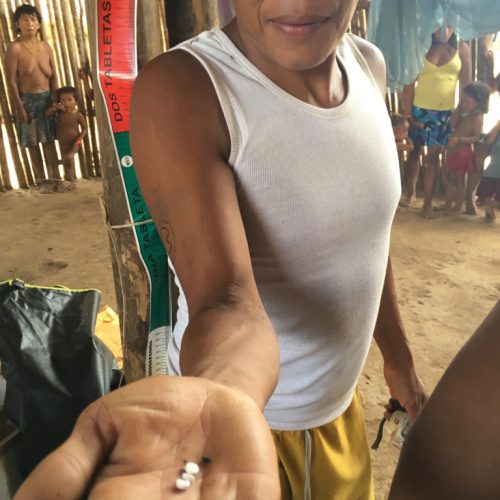 Individual displaying three small white tablets in their open hand, standing in a rustic interior with others visible in the background.